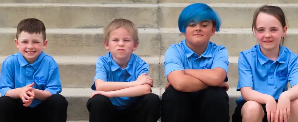 Four children sitting on outdoor stairs, wearing blue school uniforms, smiling and making different facial expressions.