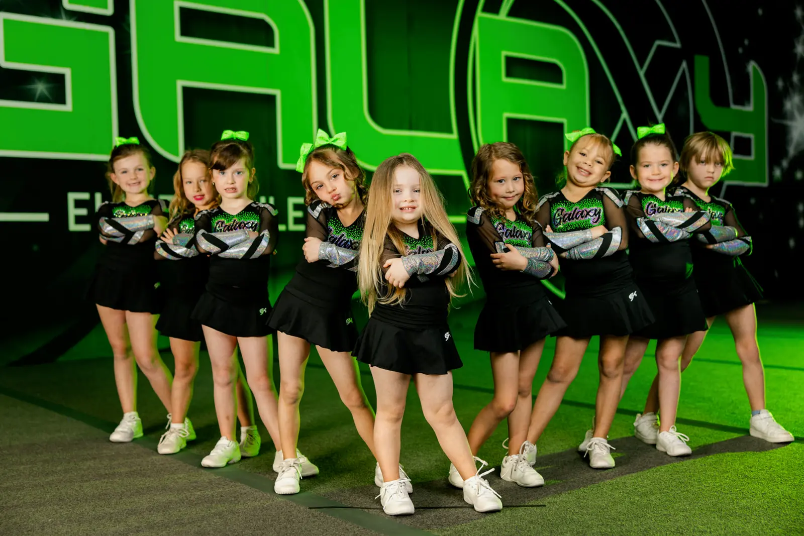 A group of young cheerleaders in uniform pose together in front of a green branded backdrop.
