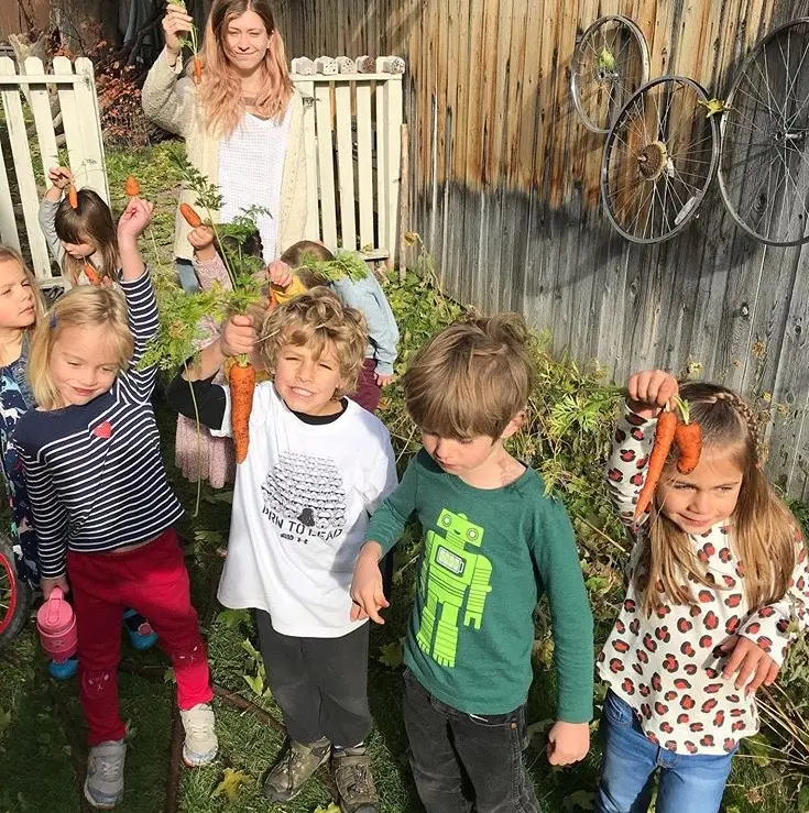 A woman and a group of children hold up freshly harvested carrots in a garden.