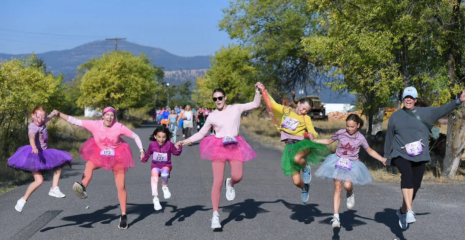A group of women and children in tutus jump together on a road during an outdoor event.
