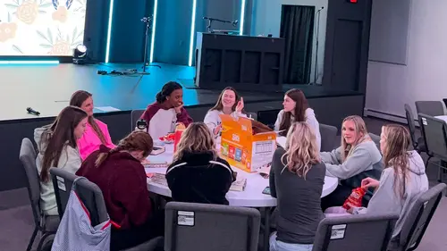 A group of young women sit around a round table in a hall with a stage in the background.