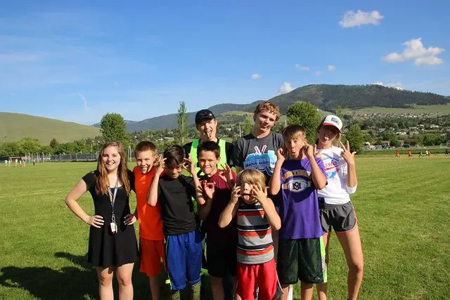 A group of young people poses outdoors on a grassy field with mountains in the background.