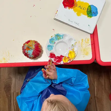 A child wearing a blue smock sits at a table painting a pumpkin with their fingers.