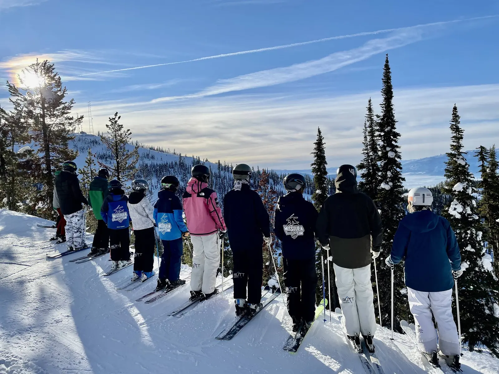 A group of skiers wearing team gear stands in a line on a snow-covered mountain ridge.