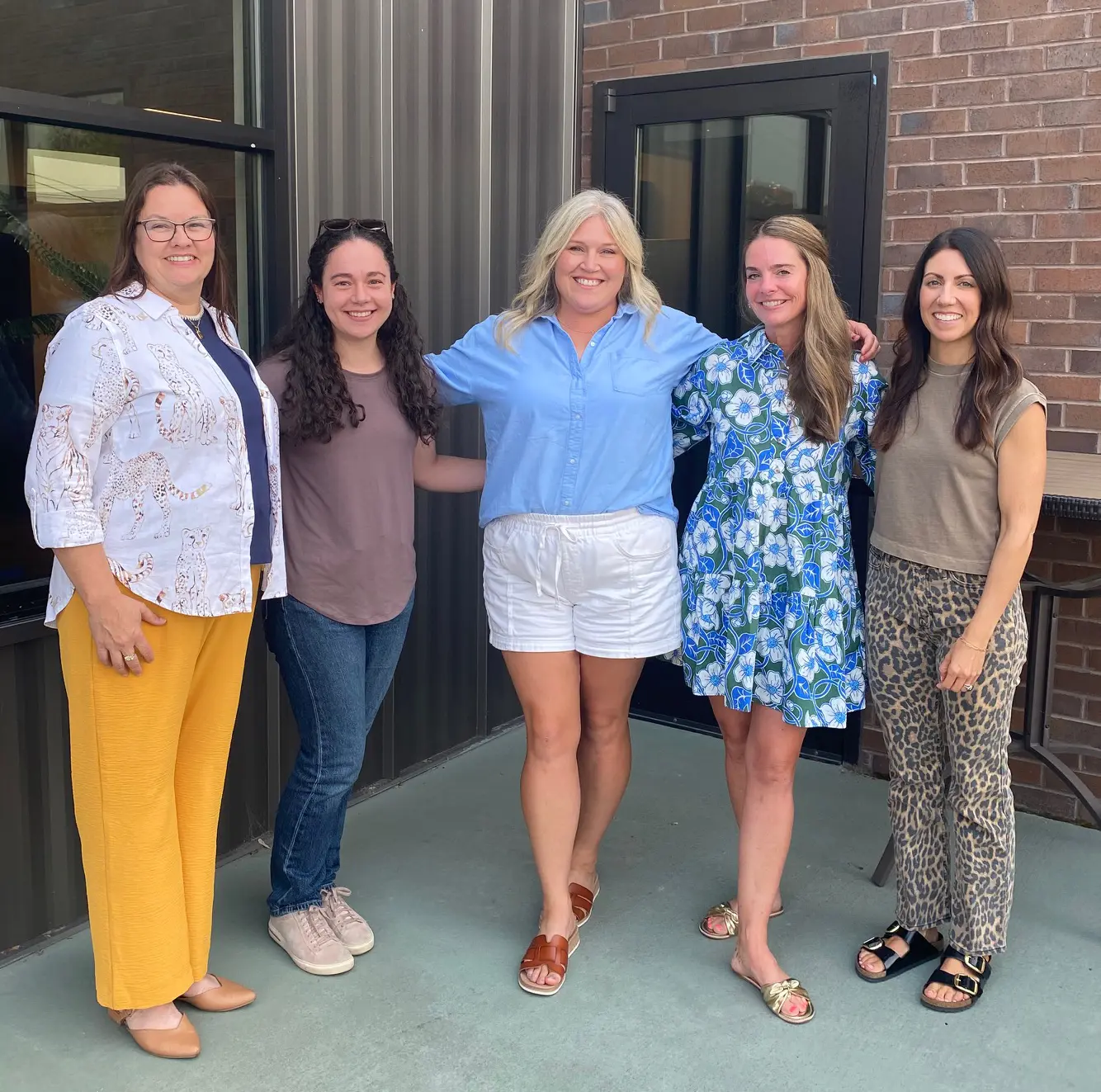 Five adult women are standing together outdoors in front of a brick building.