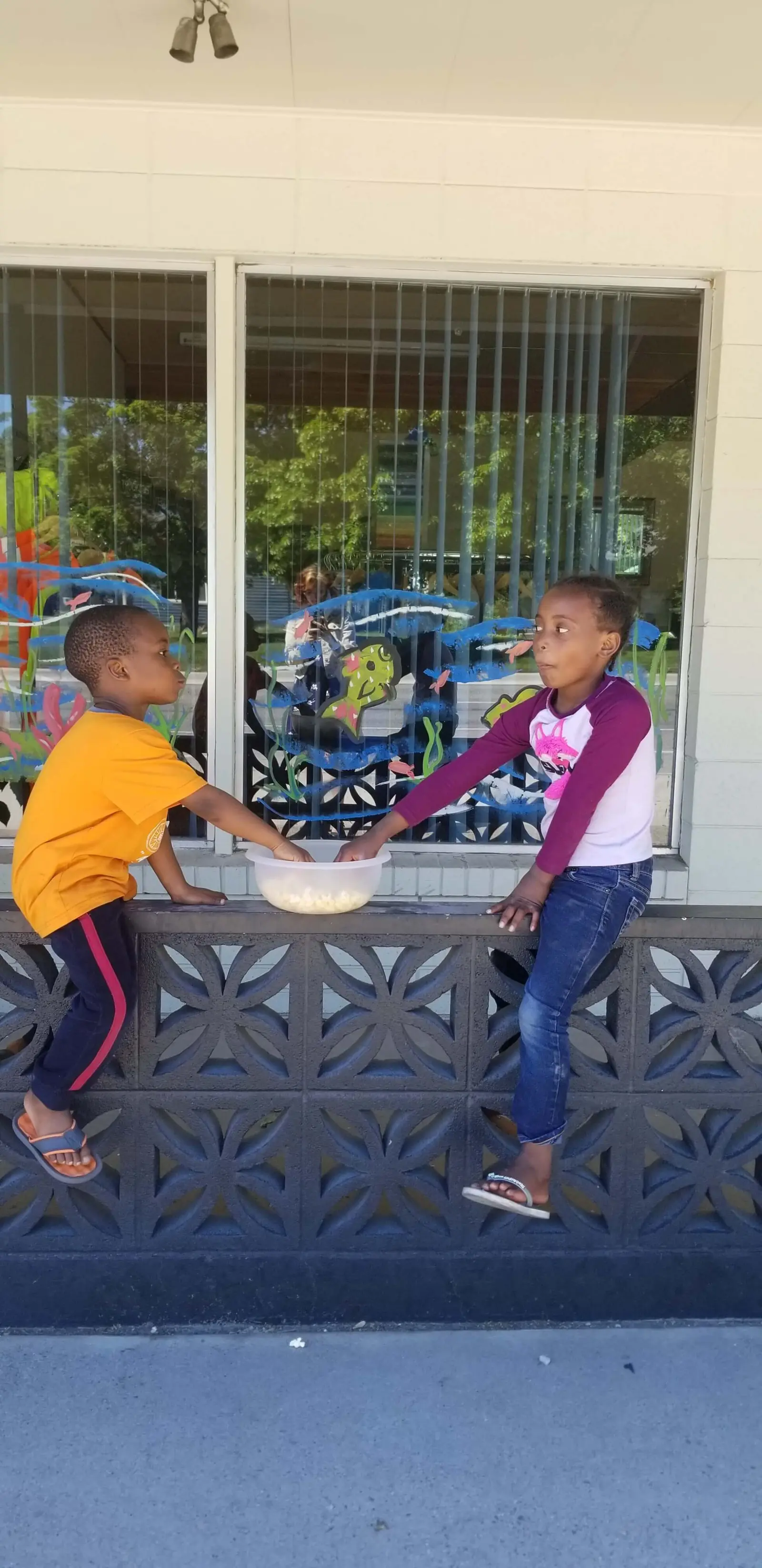 Two children sit on a concrete ledge and reach into a bowl of popcorn.