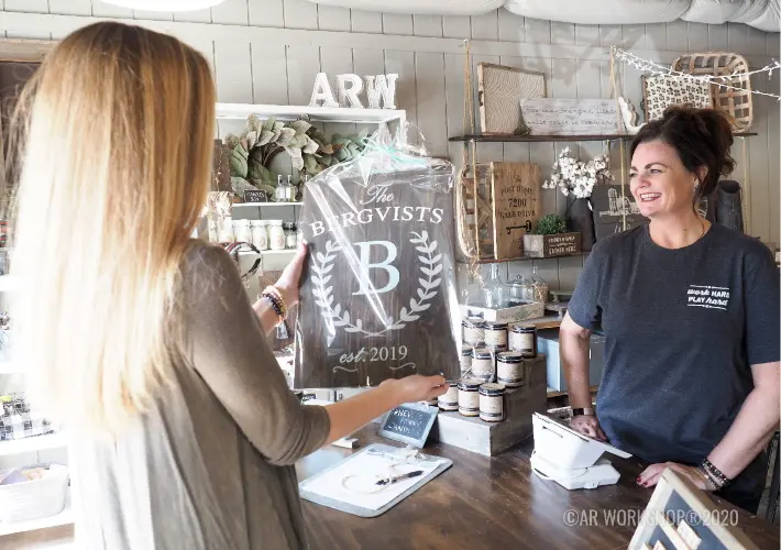 A customer hands a wooden sign to a staff member across a shop counter.