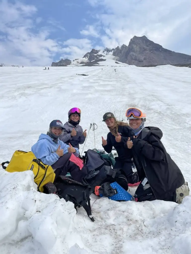 Four young skiers sit in the snow giving thumbs up on a mountain.