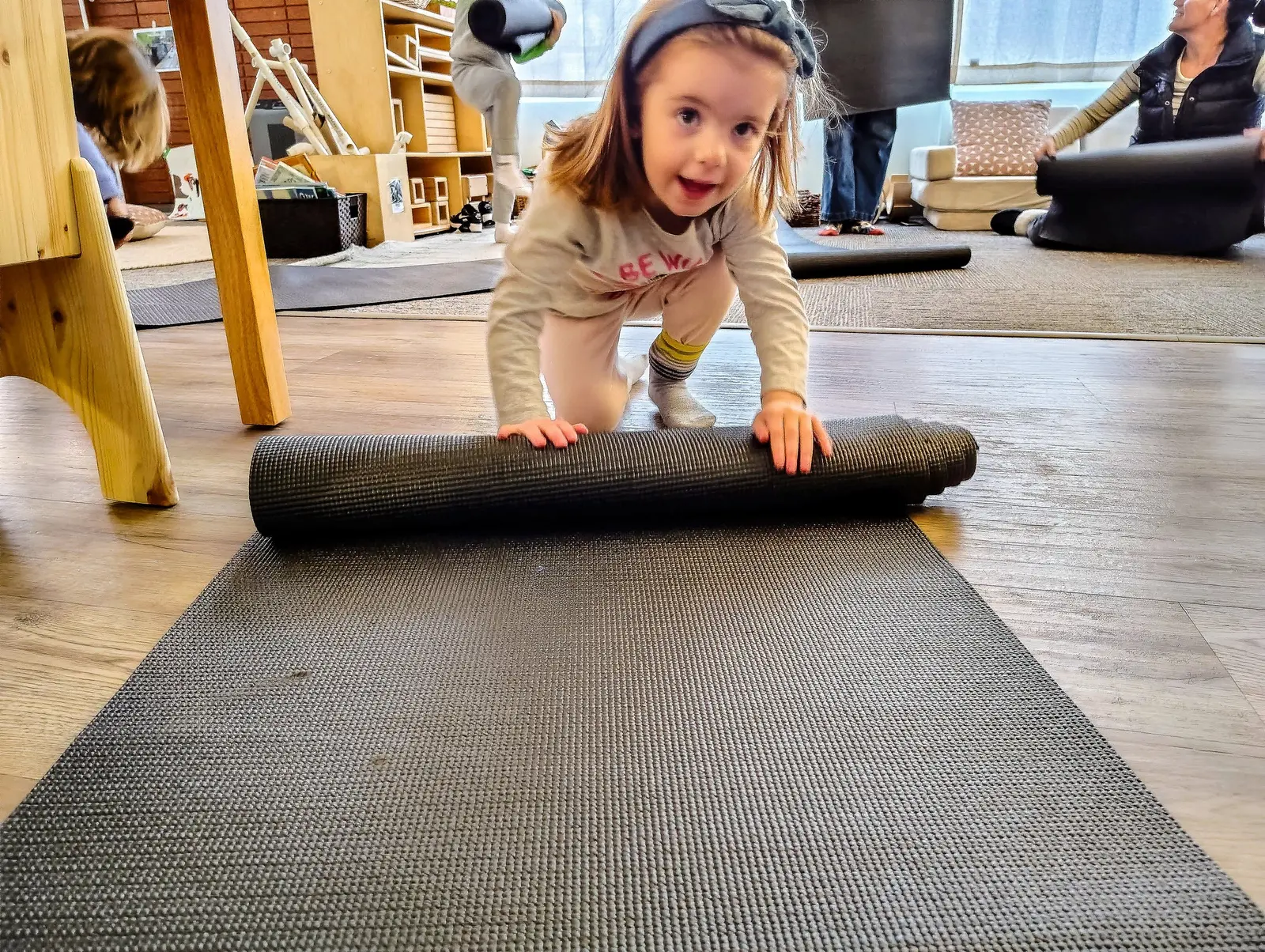 A young child rolls out a yoga mat in an indoor room with other people.