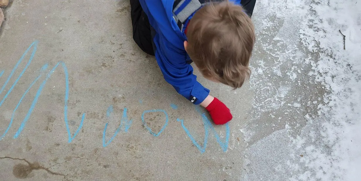 A child wearing a blue jacket and red glove draws with blue chalk on a concrete surface with snow nearby.