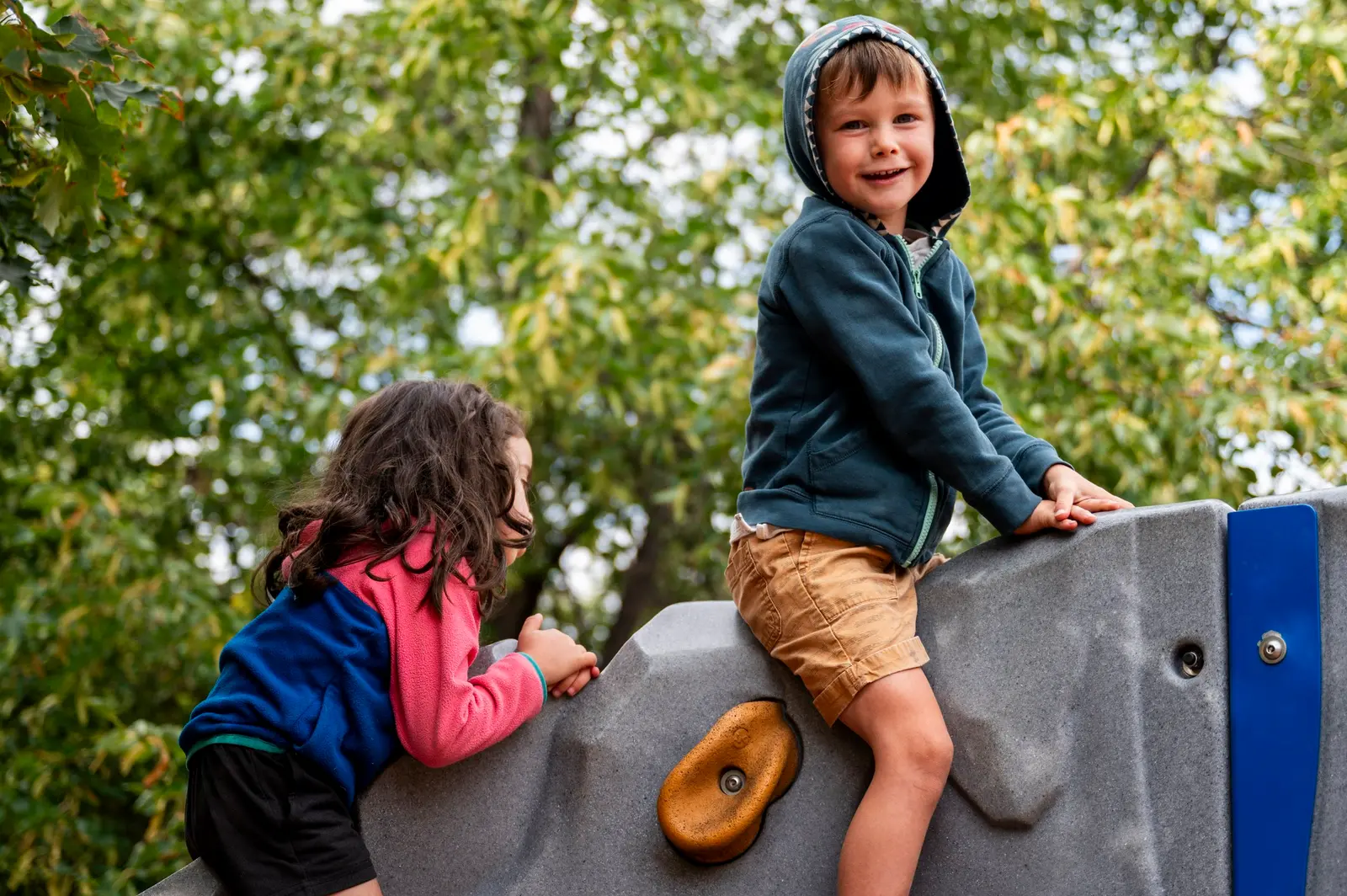 Two children climb on a rock-shaped playground feature outdoors.