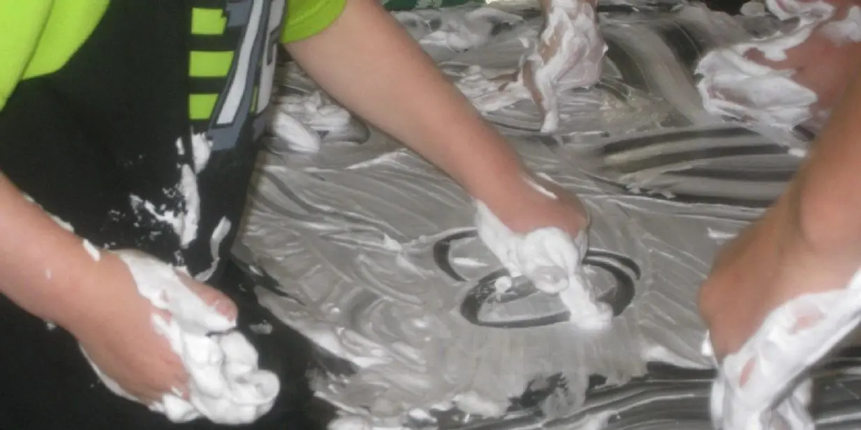 Children use their hands to trace patterns in white foam spread on a dark surface.