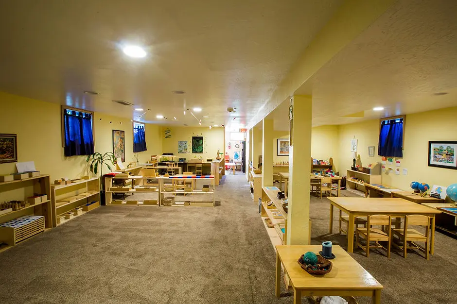 A wide view of an empty classroom arranged with low shelves containing educational materials and small wooden tables.