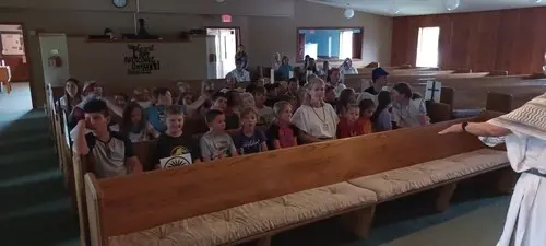 A group of children sits in church pews while an instructor leads a lesson.