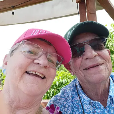 A smiling couple wearing hats and glasses takes a selfie together outdoors.