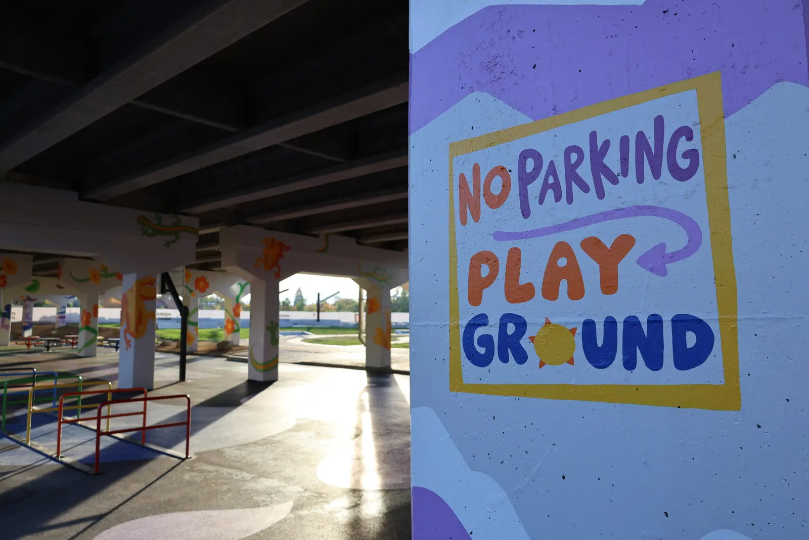 A colorful sign painted on a concrete pillar designates a playground area underneath an overpass.