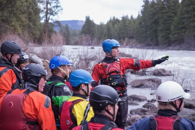 An instructor in red gear points across a river while a group of people in safety equipment watches.
