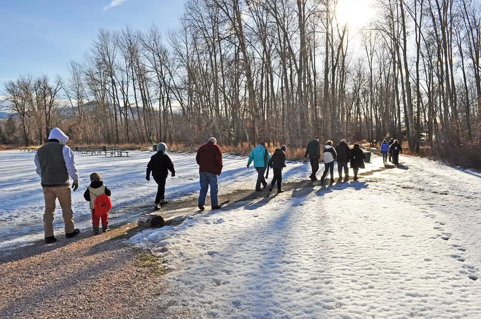A group of people walks along a snowy path through a wooded area on a sunny day.