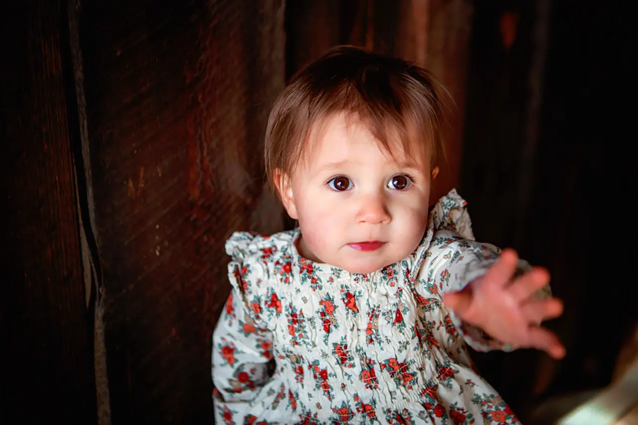 A toddler in a floral dress reaches toward the camera against a dark wooden background.