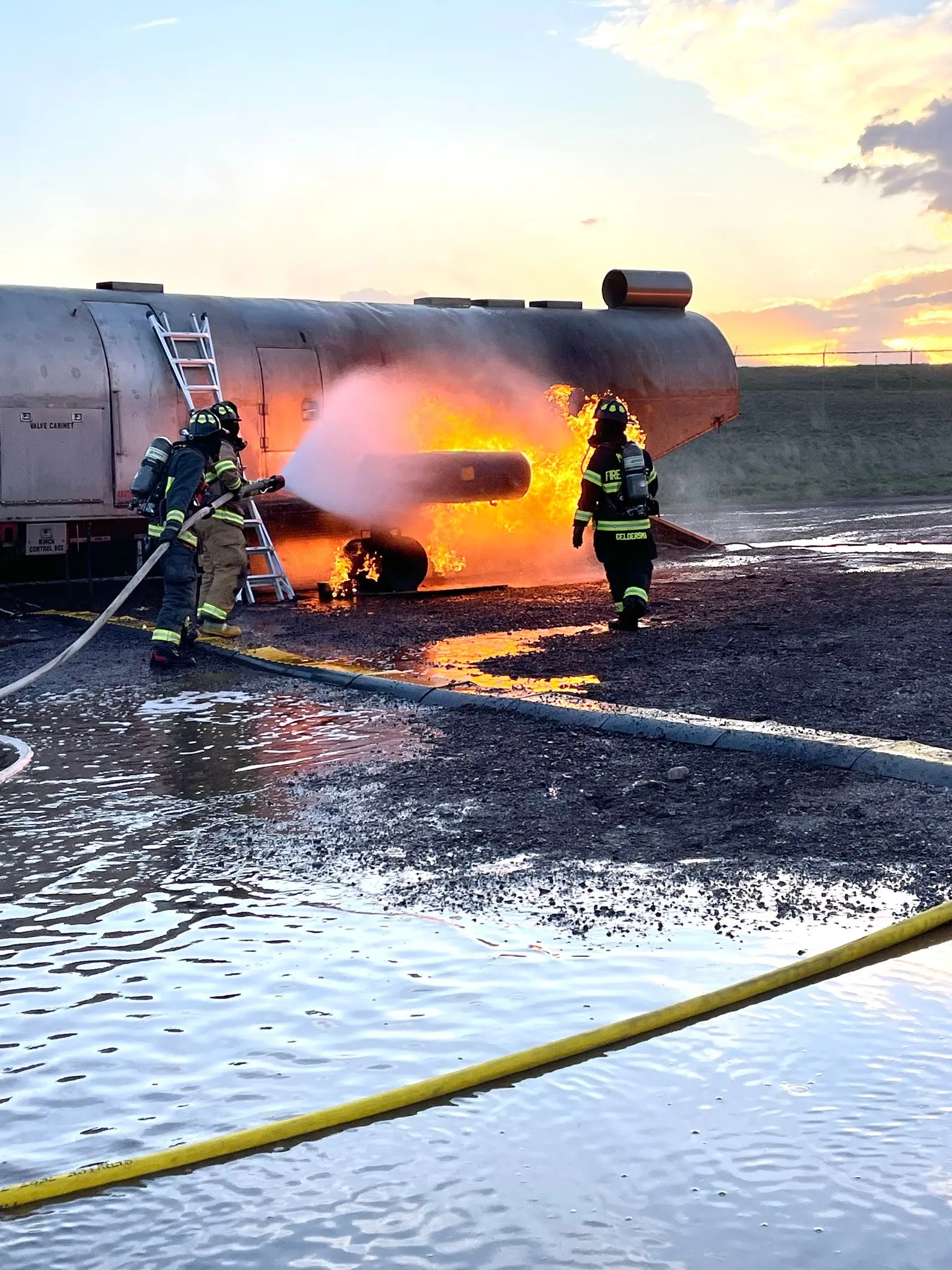 Firefighters are training by extinguishing a fire on a large metal tank container at sunset.