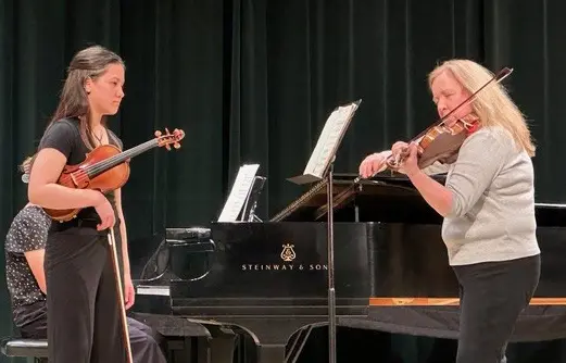 Two people play the violin while a third person sits at a grand piano.