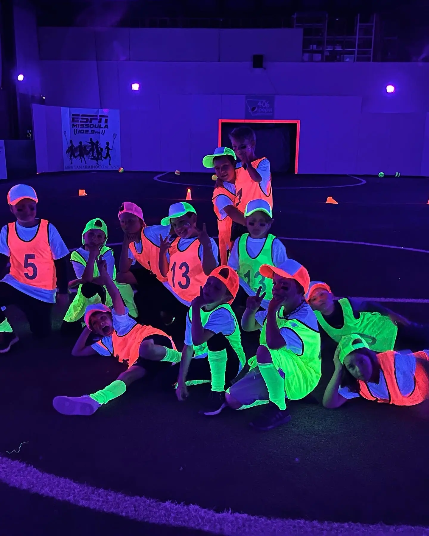 A group of children wearing neon glow-in-the-dark sports pinnies pose together in a darkened indoor arena.