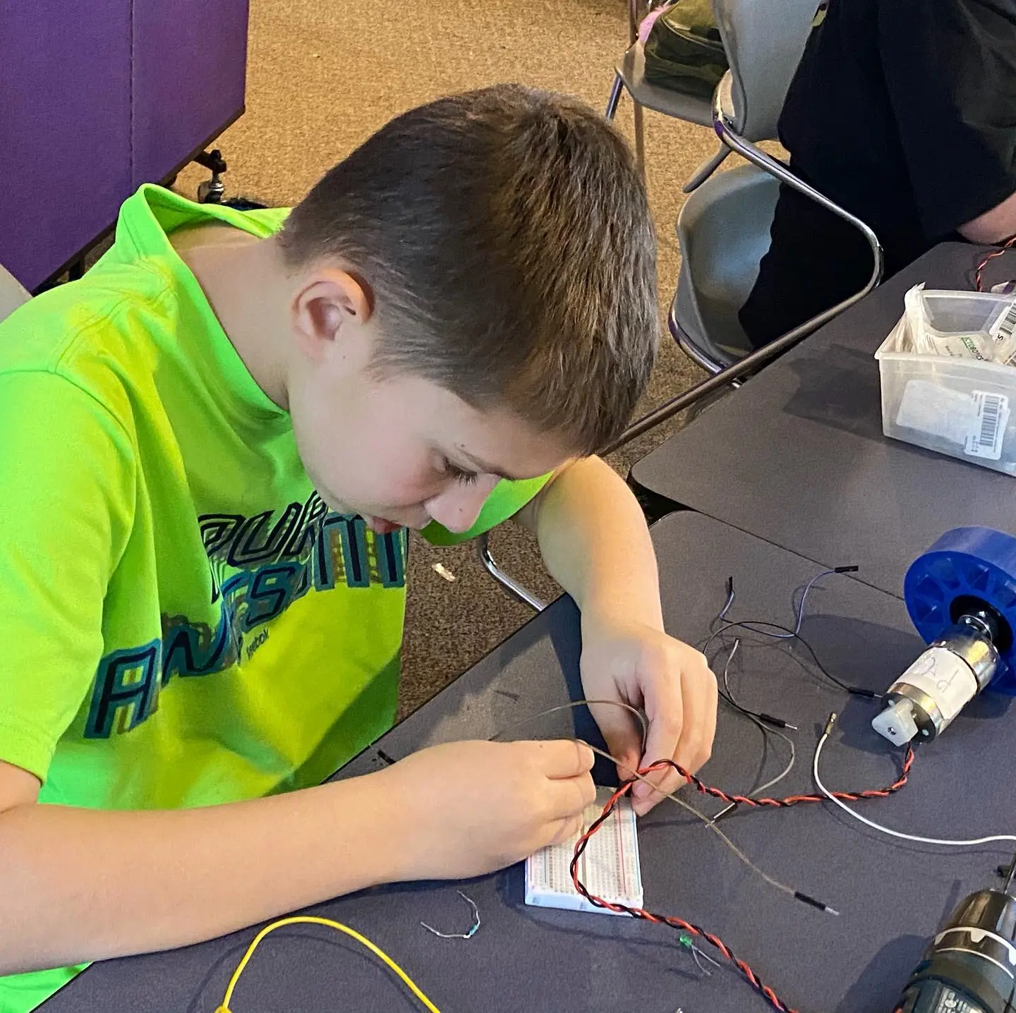 A child wearing a bright green shirt works on a robotics project at a table.