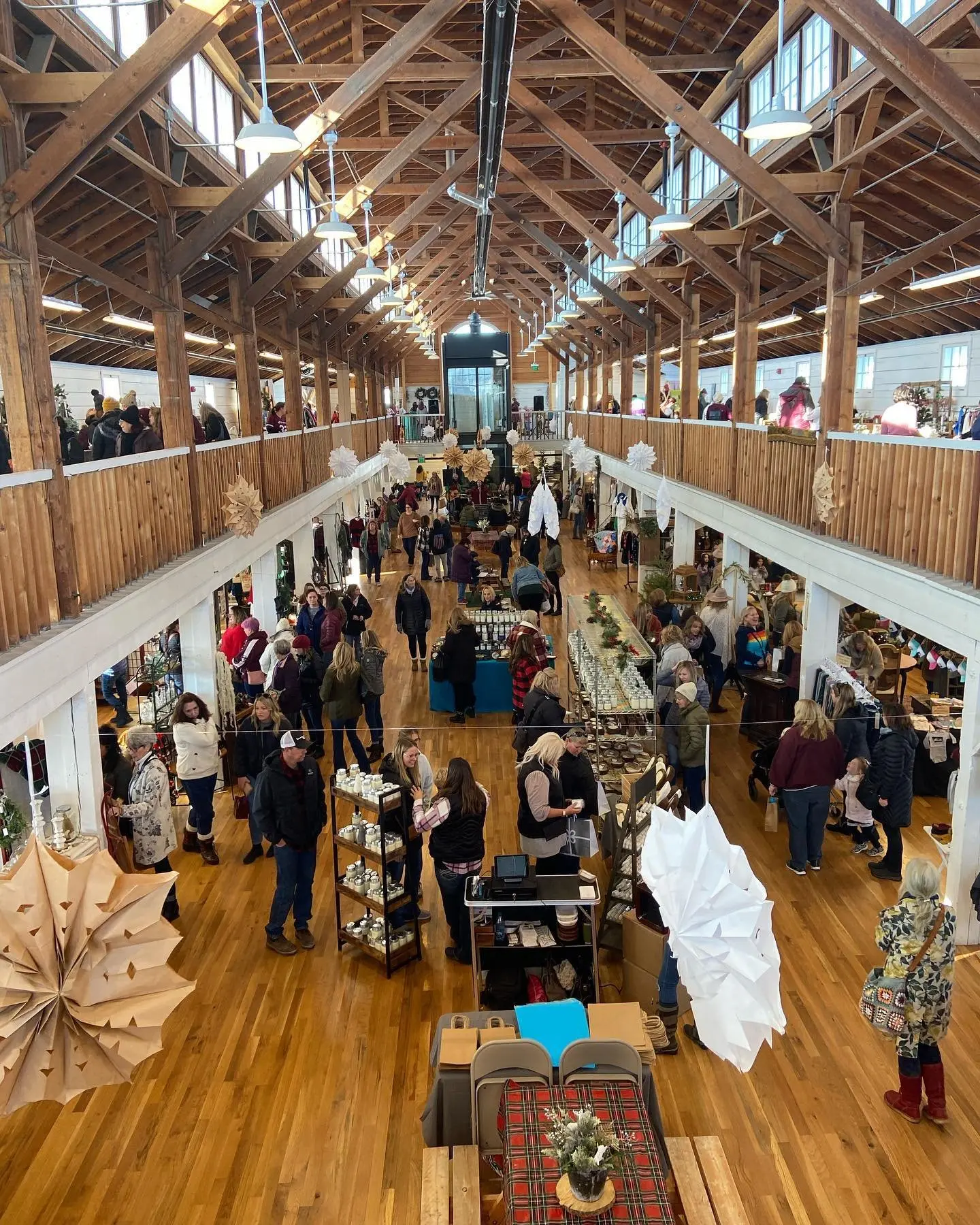 A bustling indoor market with wooden beams and high ceilings, filled with shoppers browsing various vendor booths on both the ground floor and balcony. Decorations include large paper flowers and ornaments.