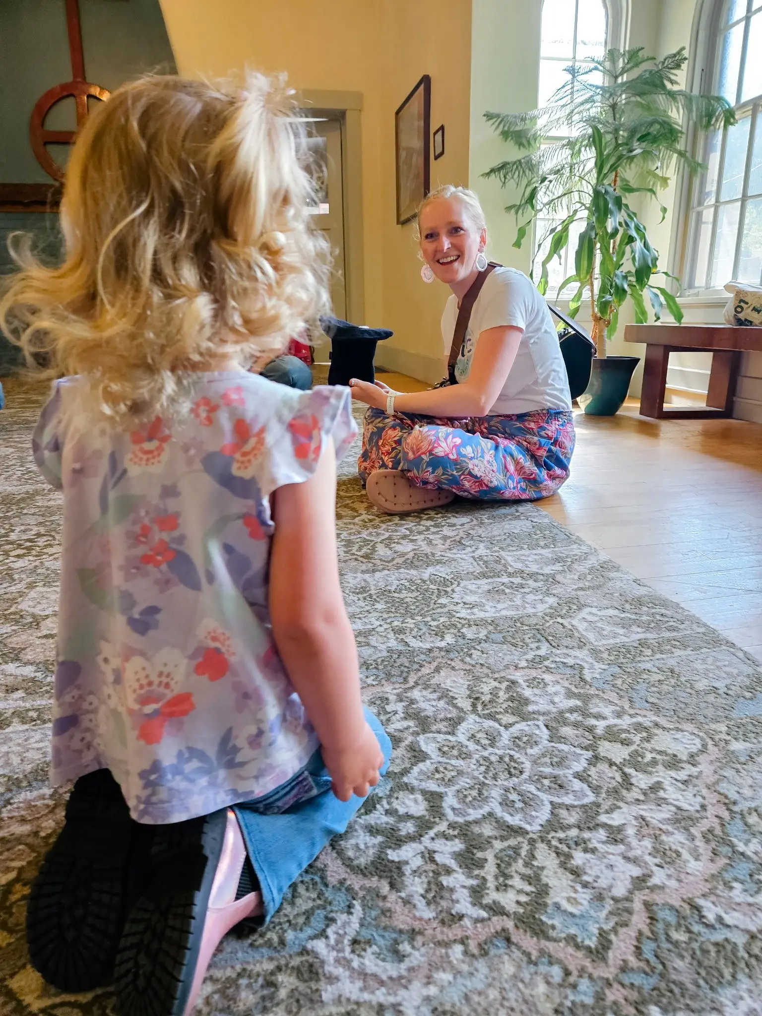 A smiling woman sits on the floor interacting with a young child.