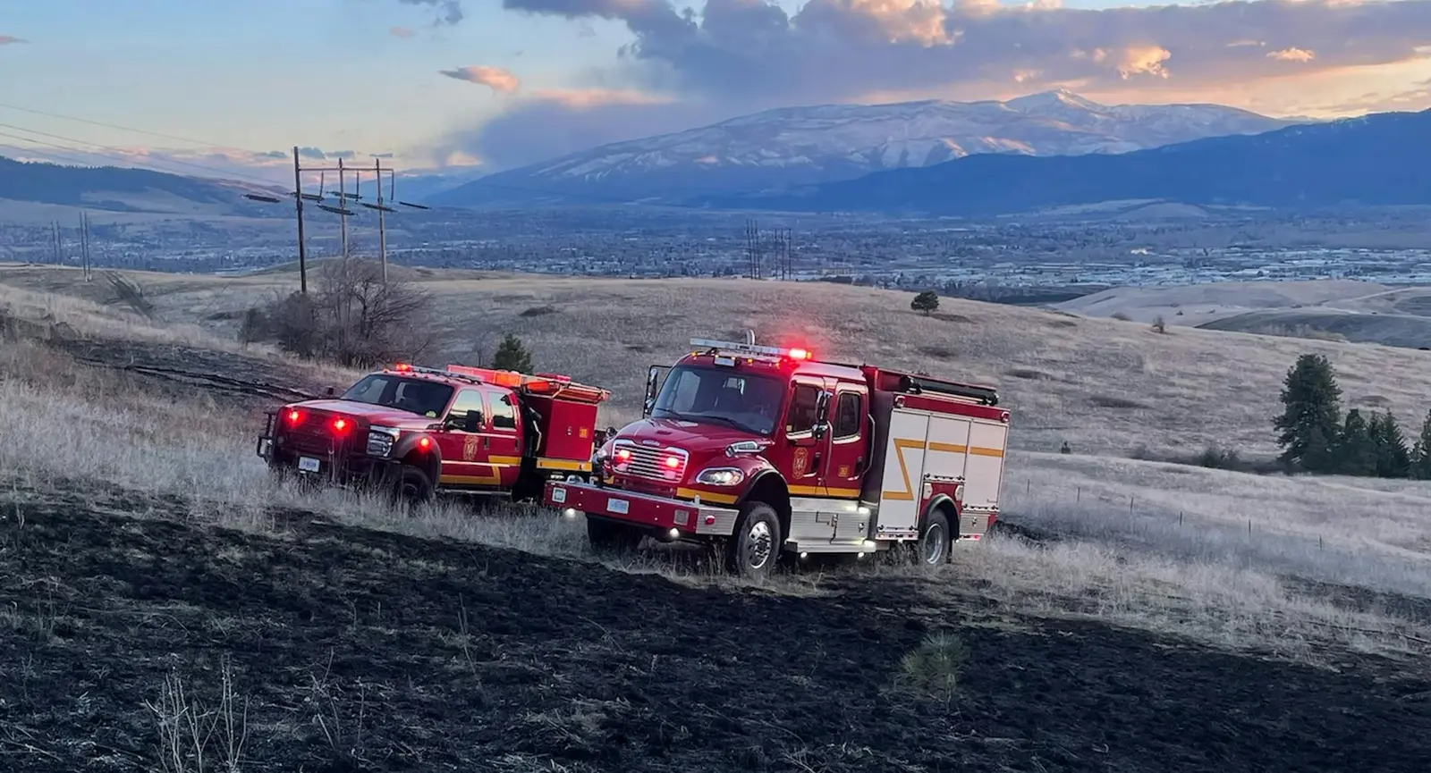 Two red fire trucks are parked on a hillside with snow-covered mountains in the background at sunset.