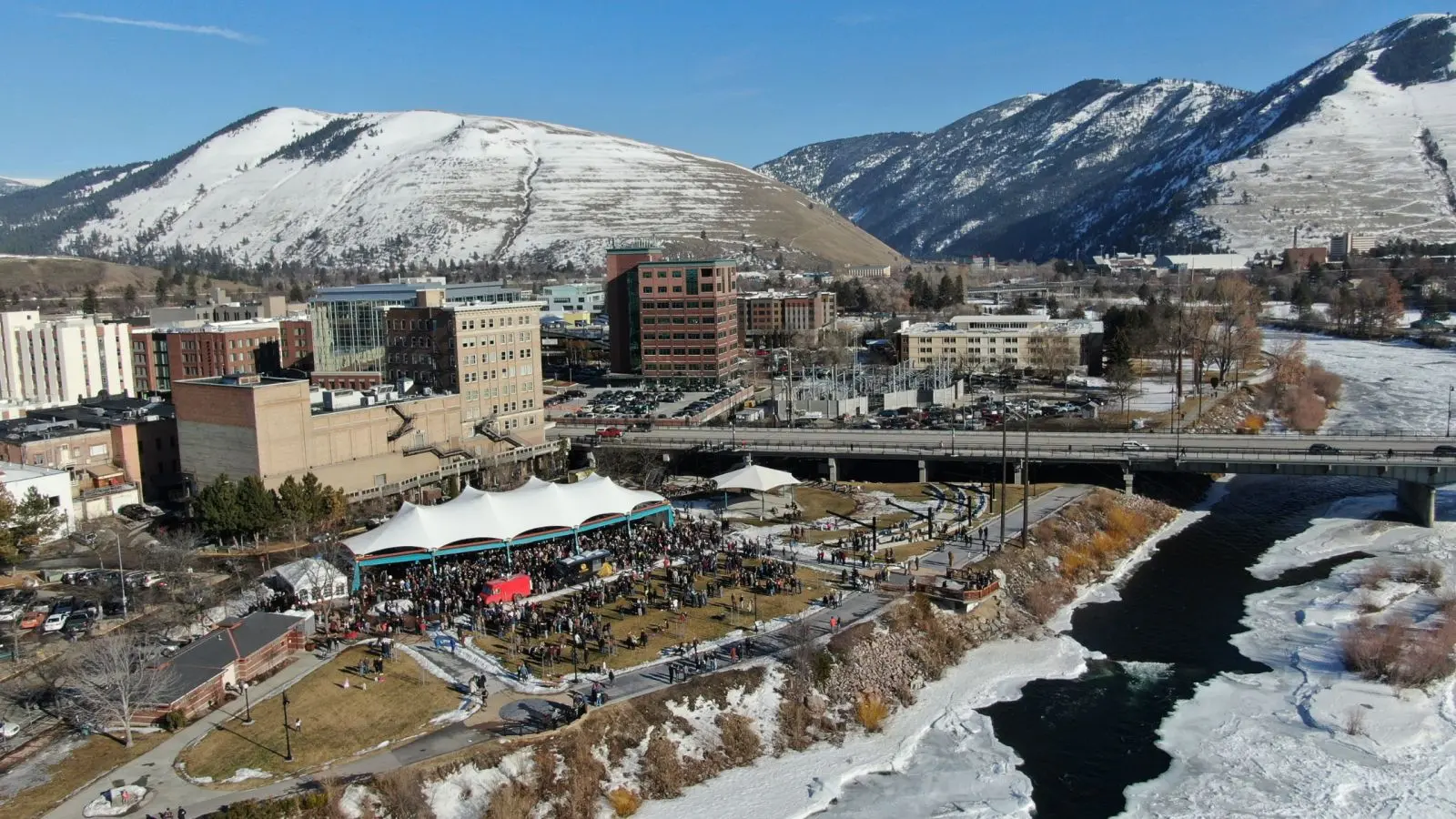 An aerial view of a town park and riverfront with snow-covered mountains in the background.