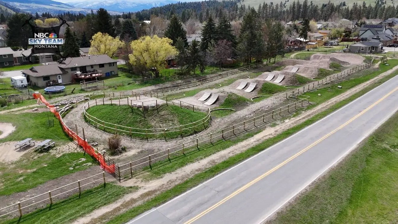 An aerial view shows a bike skills park with dirt jumps and ramps near a road and residential homes.