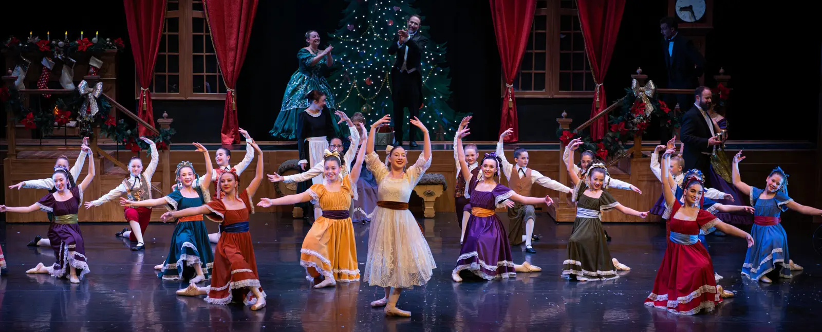 A group of dancers in colorful period-style costumes performs on a stage with a Christmas tree and holiday decor.