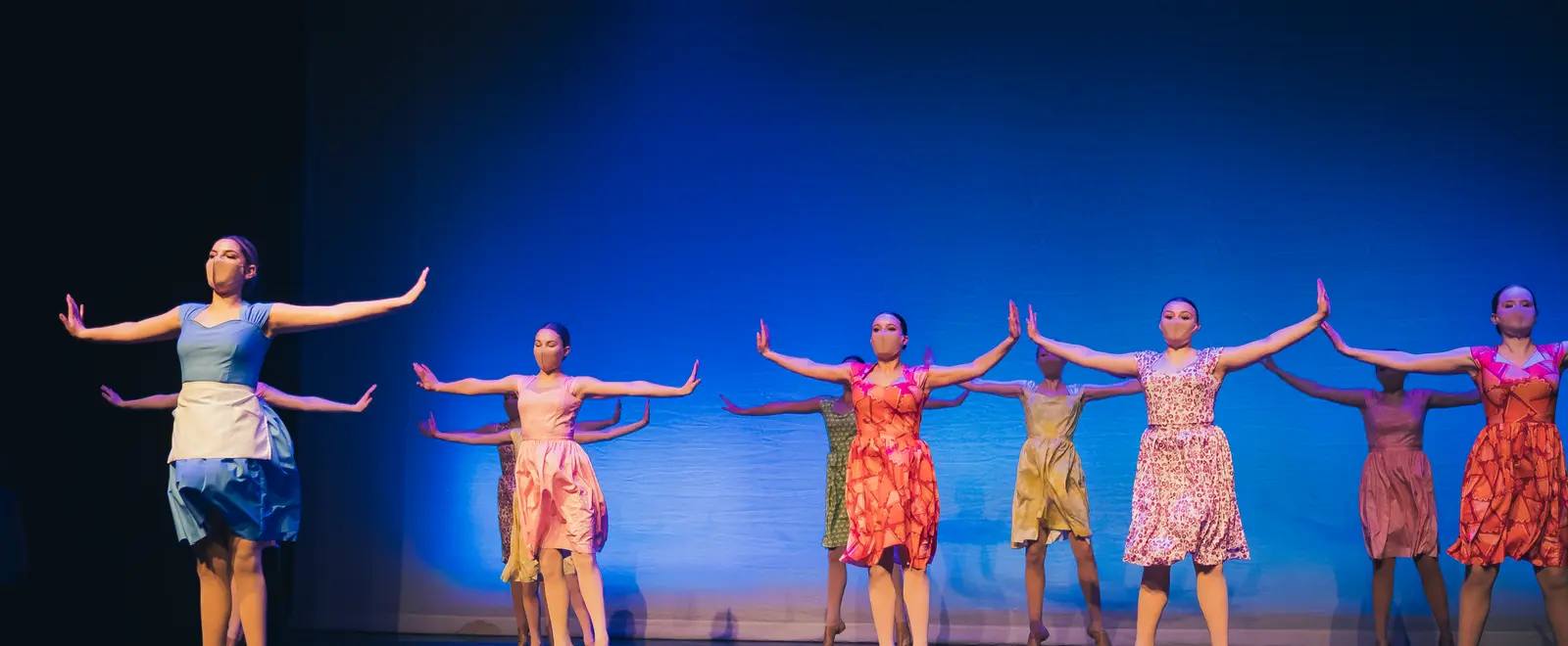 A group of dancers in colorful dresses perform on stage against a blue background.
