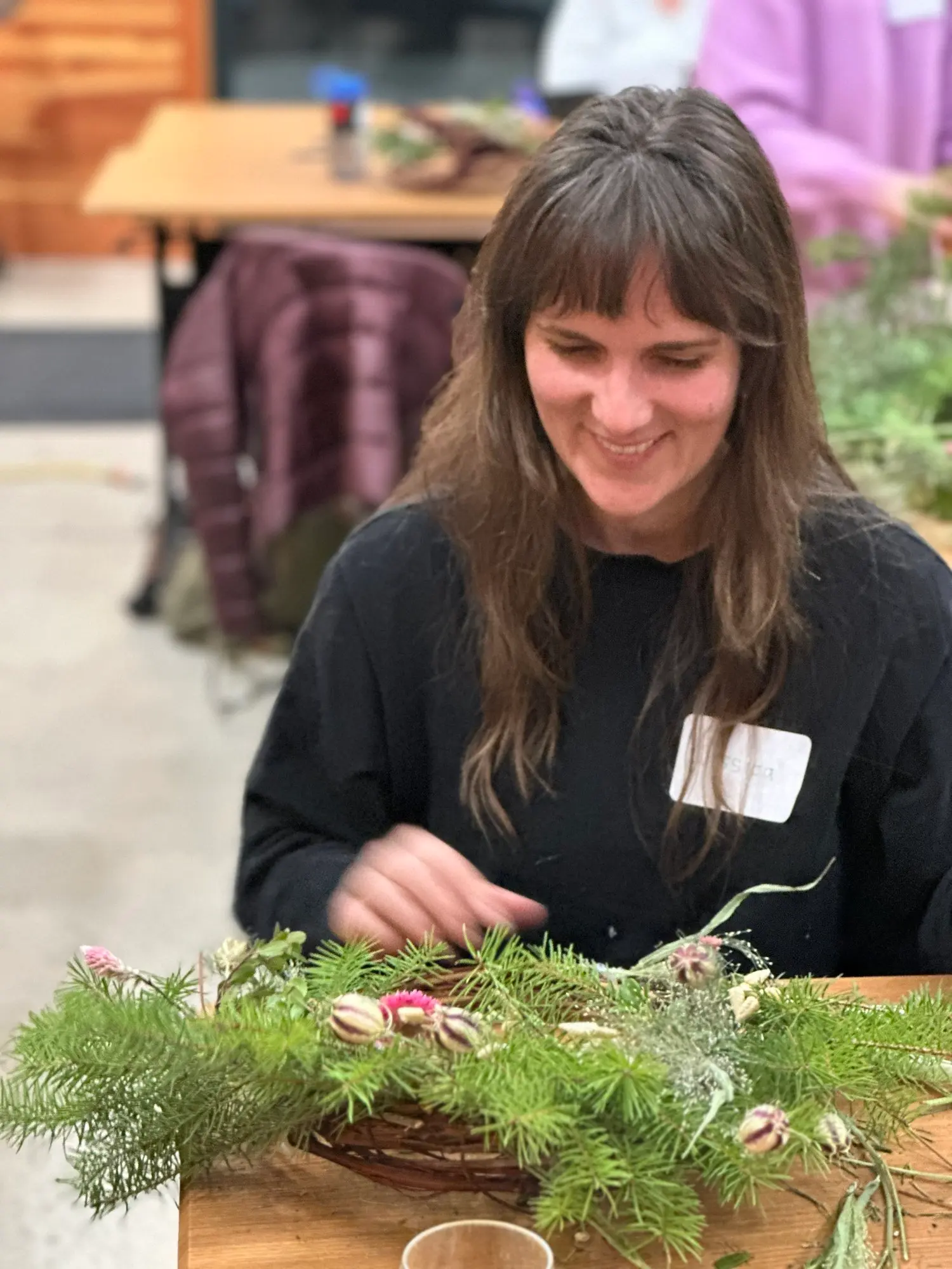 A woman smiles while crafting a wreath with evergreen branches.