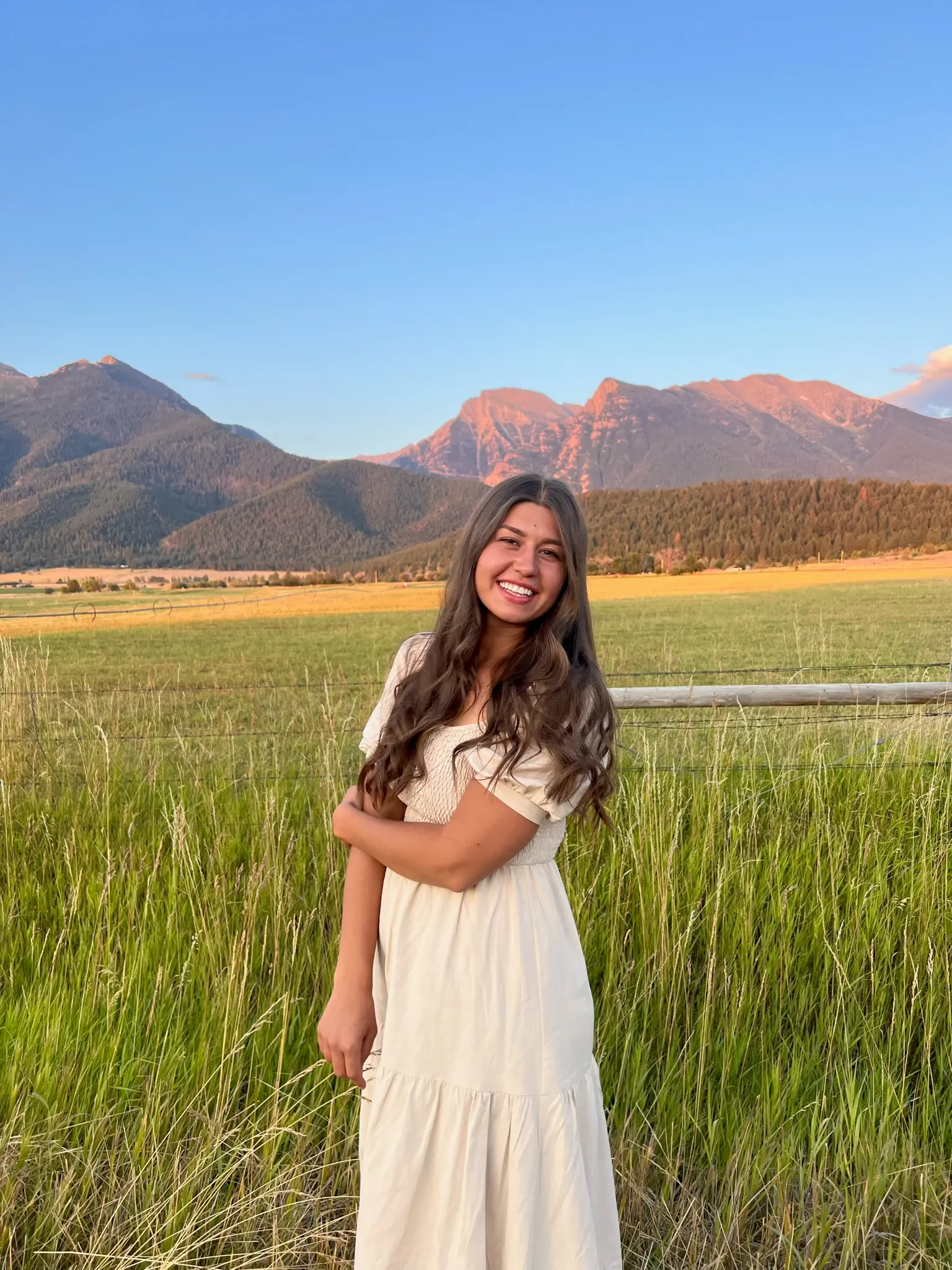 A smiling young woman stands in a grassy field in front of a mountain range at sunset.