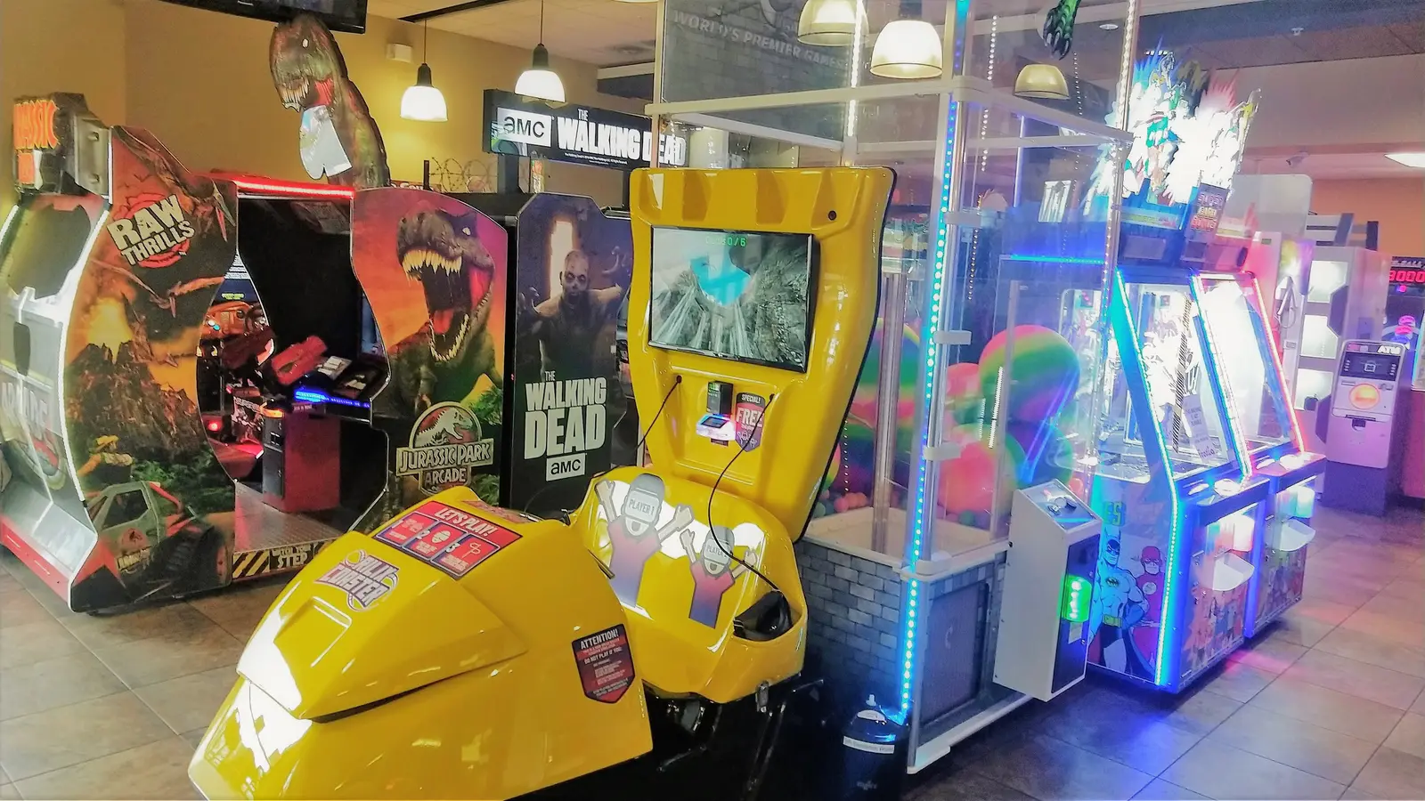 An indoor arcade features several video game cabinets including a yellow motion simulator and a Jurassic Park game.