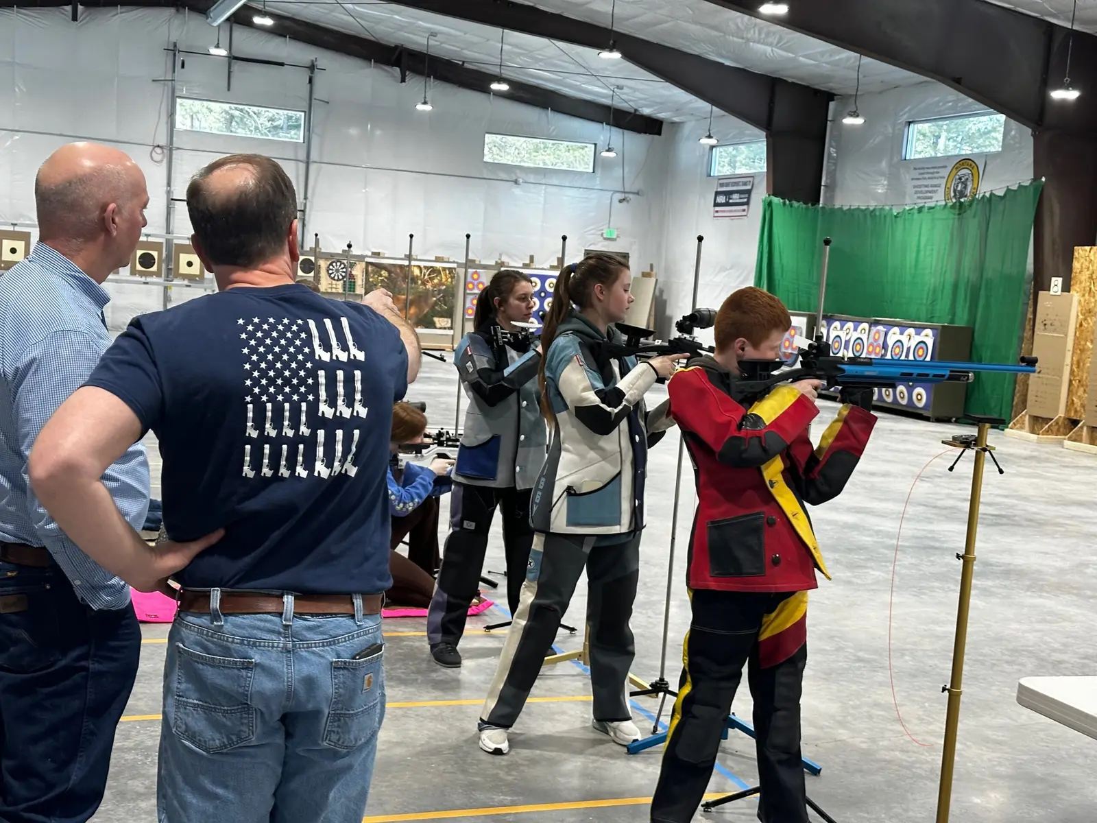 Adult instructors watch youths practice shooting at an indoor range.