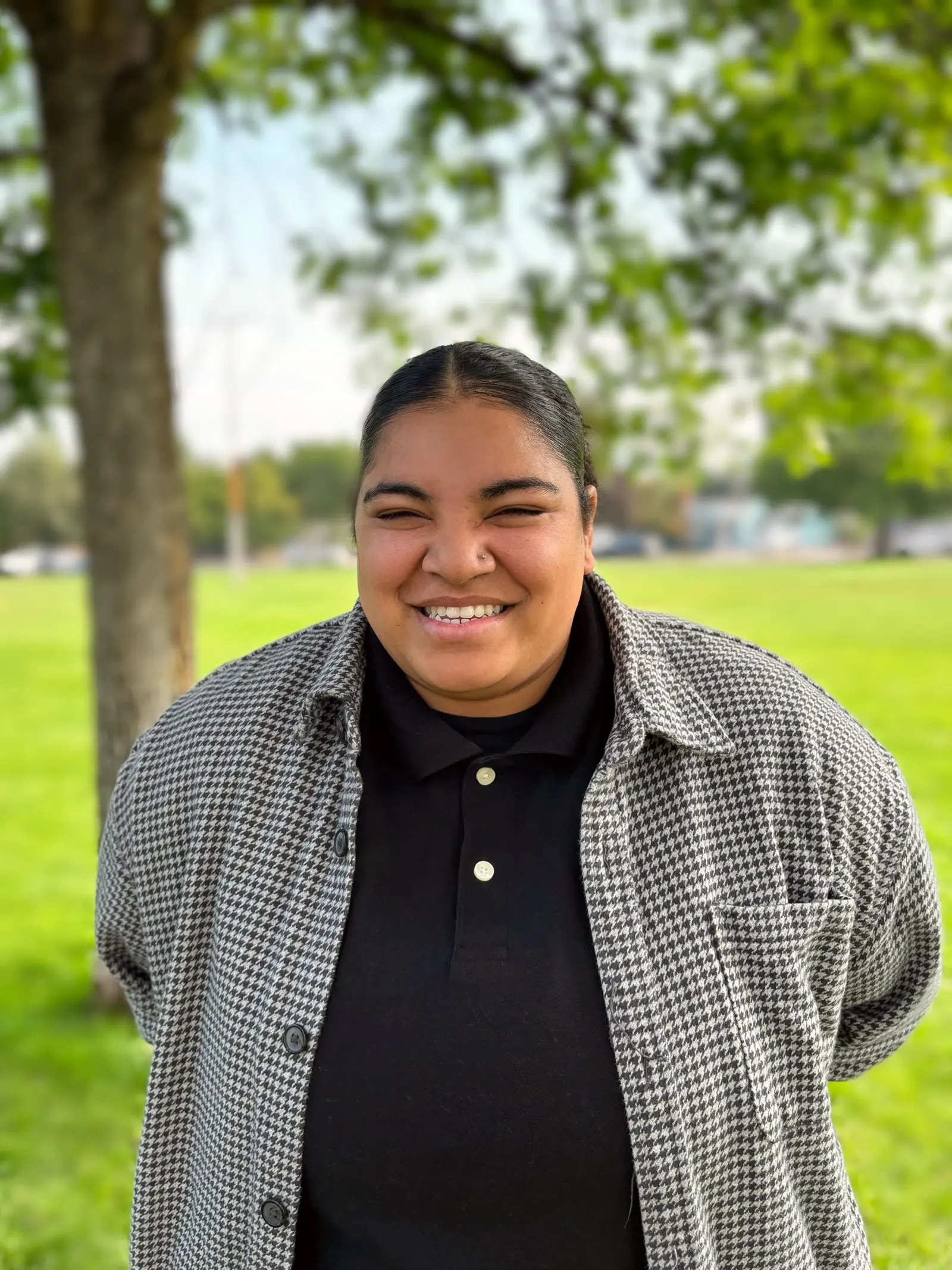 A smiling person stands outside in a park.