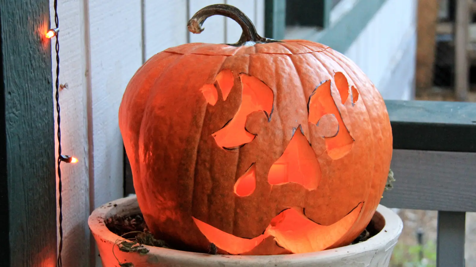 A lit jack-o'-lantern sits in a white pot on a porch.