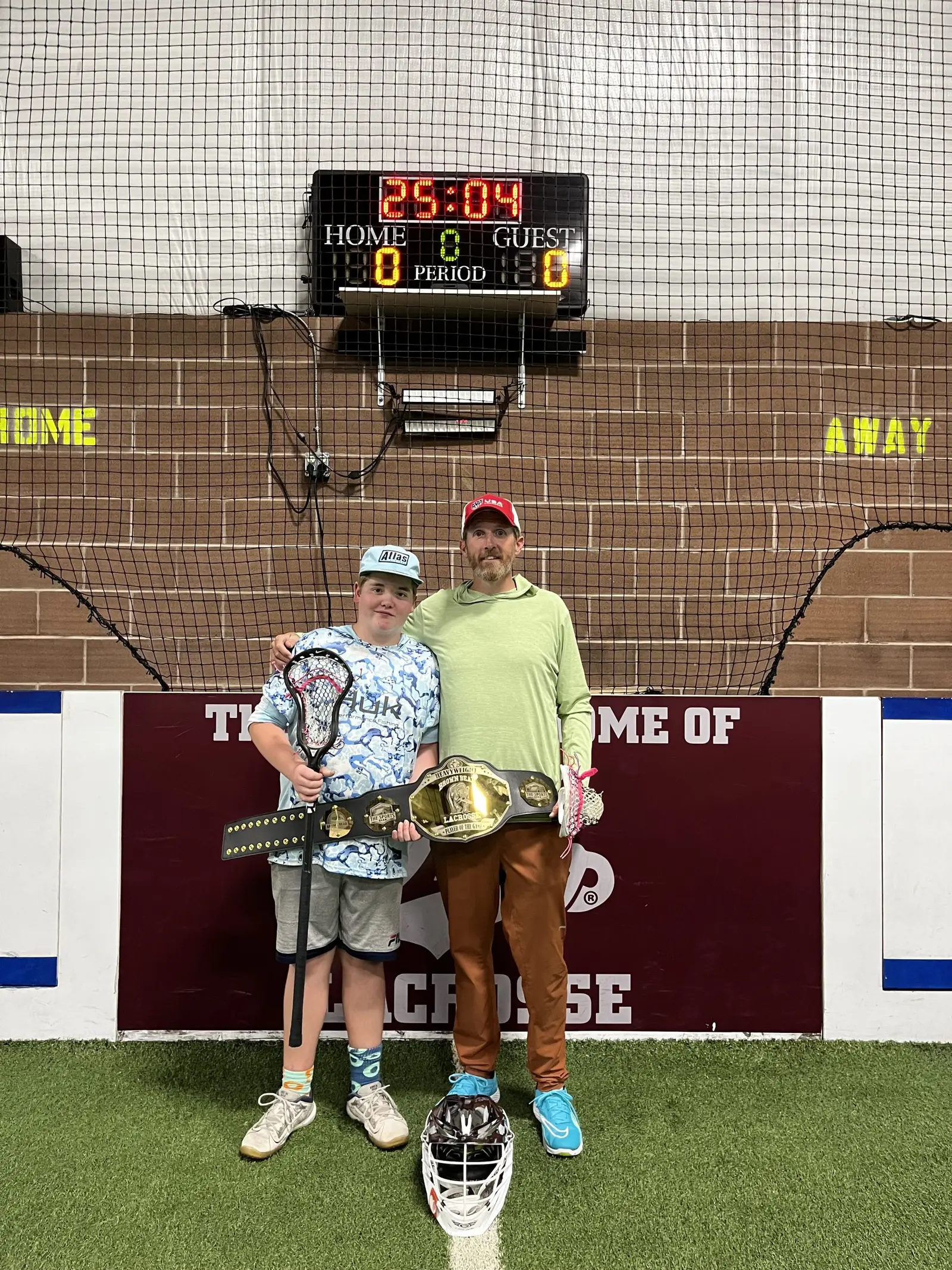A coach and a player stand together holding a championship belt in an indoor sports facility.