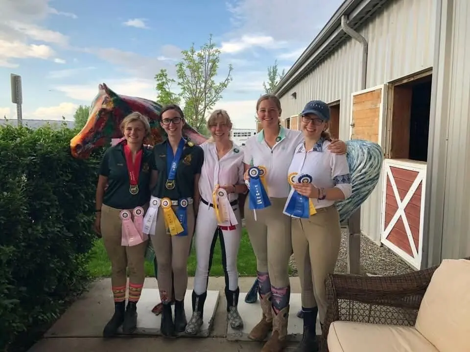 Five young women standing together wearing equestrian attire and ribbons.