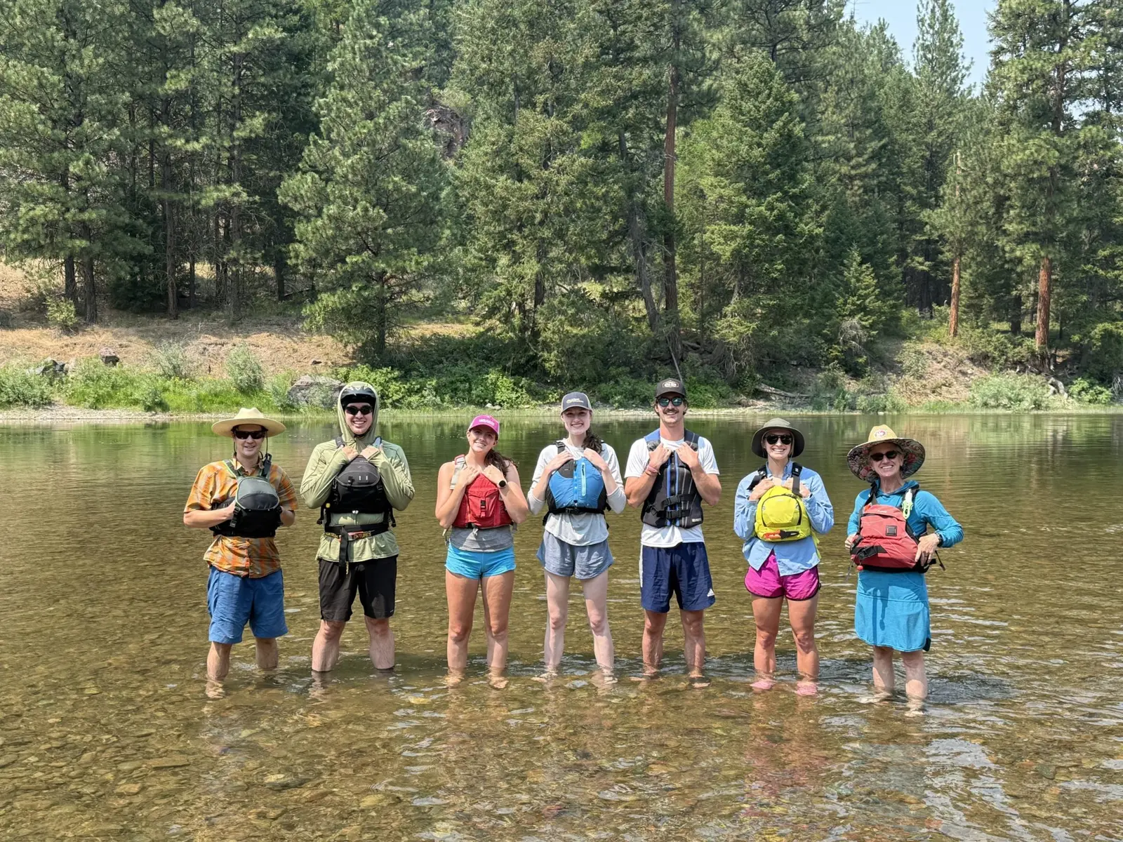 A group of seven adults wearing life jackets stand in a river in front of a forest.