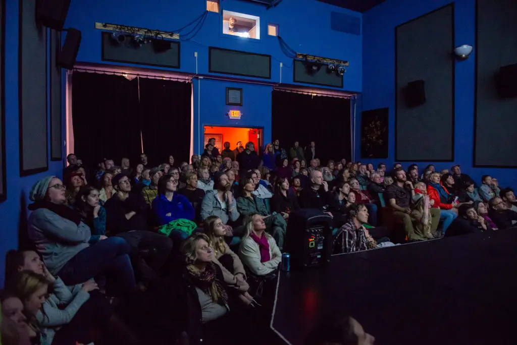 A crowd of people sits in a theater watching a performance or screening.