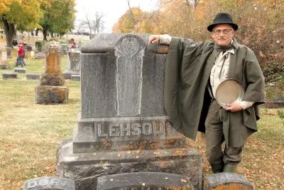 A man wearing a hat and cloak stands next to a tombstone in a cemetery.