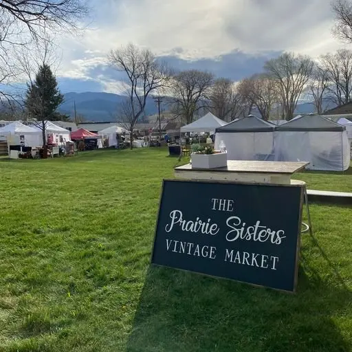 Outdoor scene of The Prairie Sisters Vintage Market with tents and market stalls on a grassy field, some trees, and mountains in the background on a partly cloudy day.