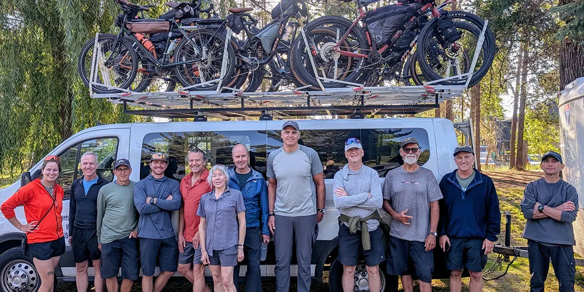 A group of adults stands in front of a van with mountain bikes mounted on a roof rack.