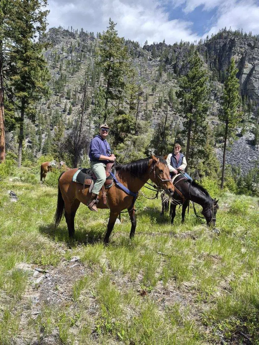 Two people ride horses on a grassy hillside beneath a mountain.