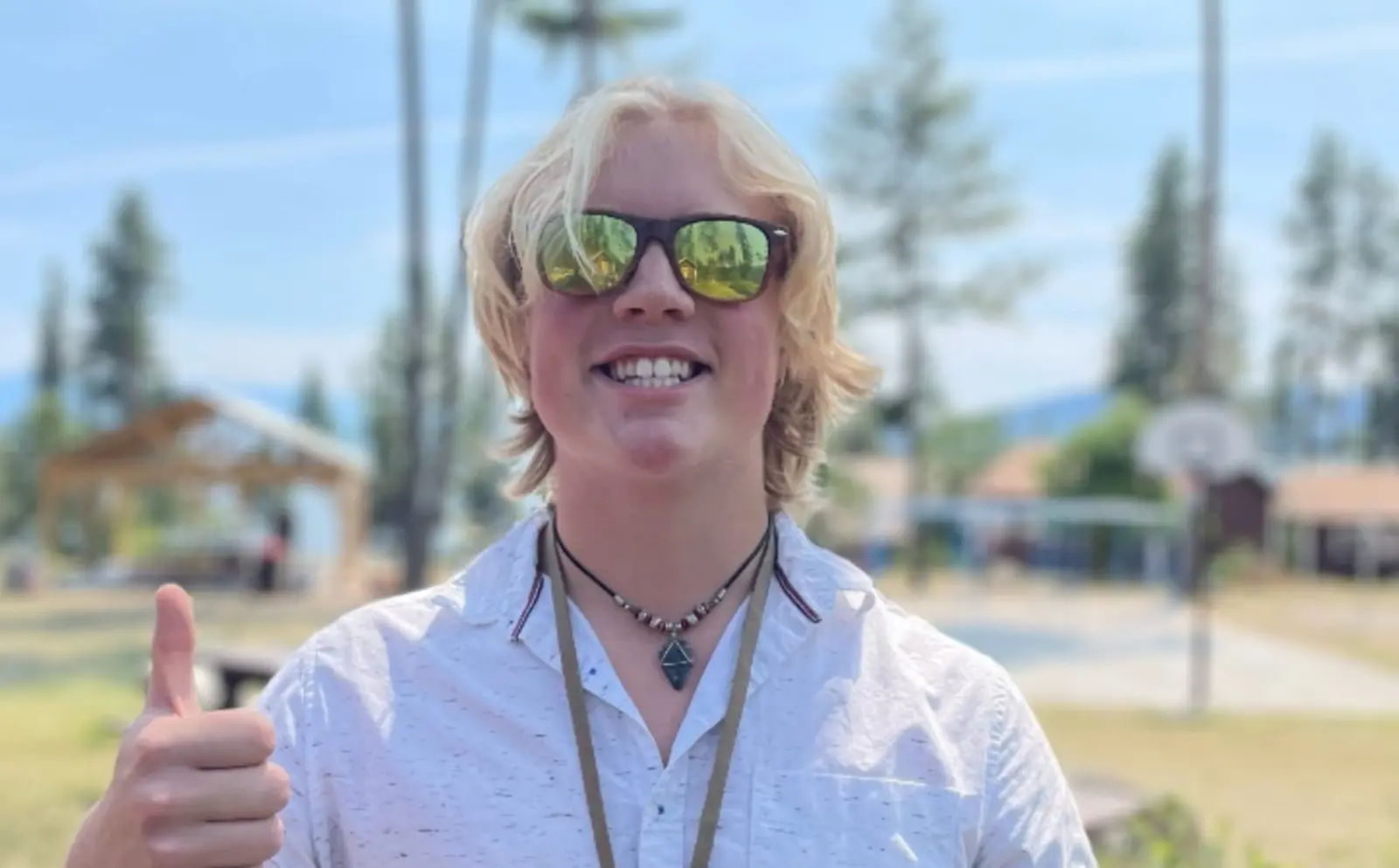 A smiling person with blonde hair and sunglasses gives a thumbs-up outdoors.