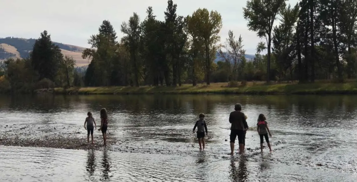 A group of children and an adult stand in shallow water by a riverbank.
